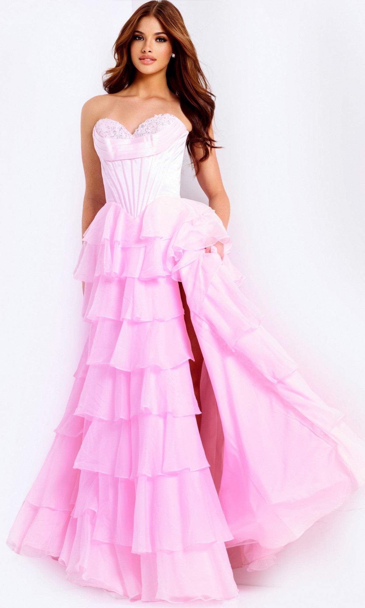 A woman poses against a white background in the Jovani 47762 long prom dress, featuring a strapless satin corset bodice and a layered light pink ruffle-tiered chiffon skirt she lifts gracefully.